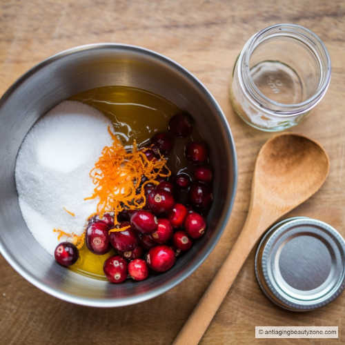 Photo of a bowl containing a mixture of granulated sugar, melted coconut oil, a small amount of mashed cranberries, and zest from an orange. A wooden spoon is next to the bowl and a small empty mason jar and lid.